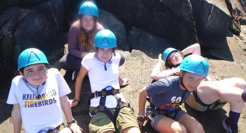 A group of students wearing blue helmets and climbing harnesses take a rest on the ground.  